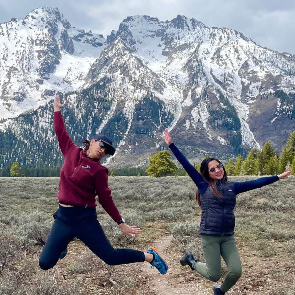 Two young ladies jump and smile in front of the snow covered Grand Teton mountains. Taken on a Teton Excursions tour of Grand Teton National Park.