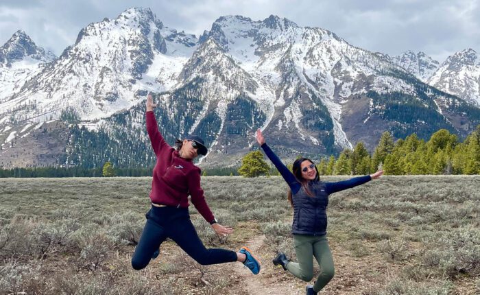 Two young ladies jump and smile in front of the snow covered Grand Teton mountains. Taken on a Teton Excursions tour of Grand Teton National Park.