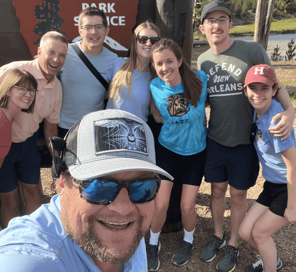 A young family and guide pose in front of the sign for the south entrance of Yellowstone National Park. Taken on a Teton Excursions tour to Yellowstone.