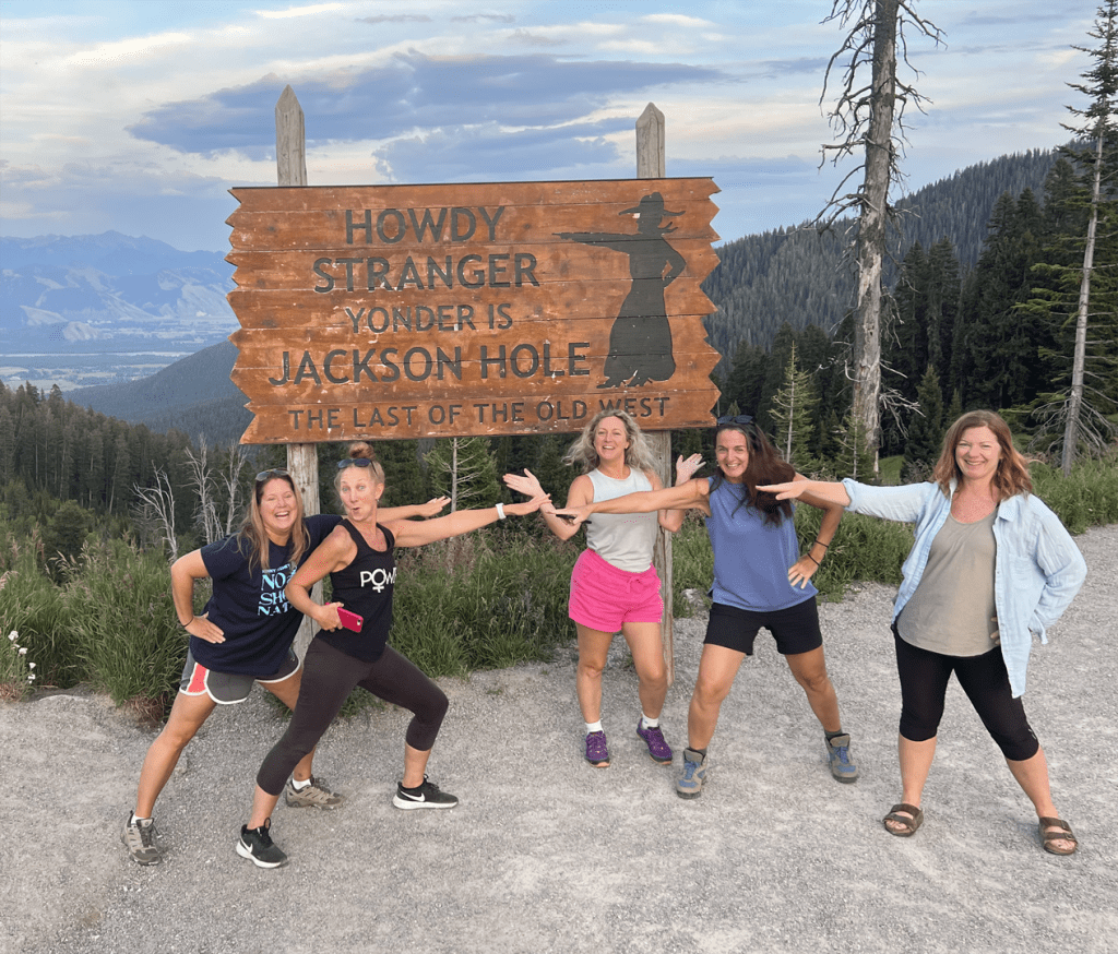 A group of young ladies poses at Teton Pass, WY in front of the sign 'Howdy Stranger, Yonder is Jackson Hole The Last of the Old West' taken on a Teton Excursions tour to Yellowstone National Park