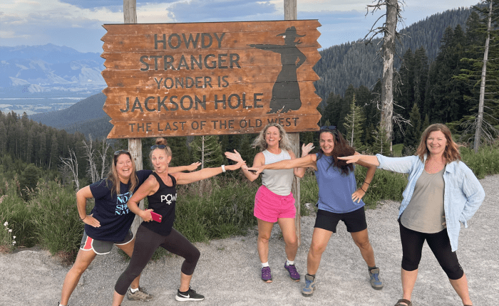 A group of young ladies poses at Teton Pass, WY in front of the sign 'Howdy Stranger, Yonder is Jackson Hole The Last of the Old West' taken on a Teton Excursions tour to Yellowstone National Park