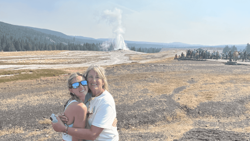 A mother and daughter pose together for a photo as Old Faithful geyser erupts in the background. Taken on a Teton Excursions tour of Yellowstone National Park.
