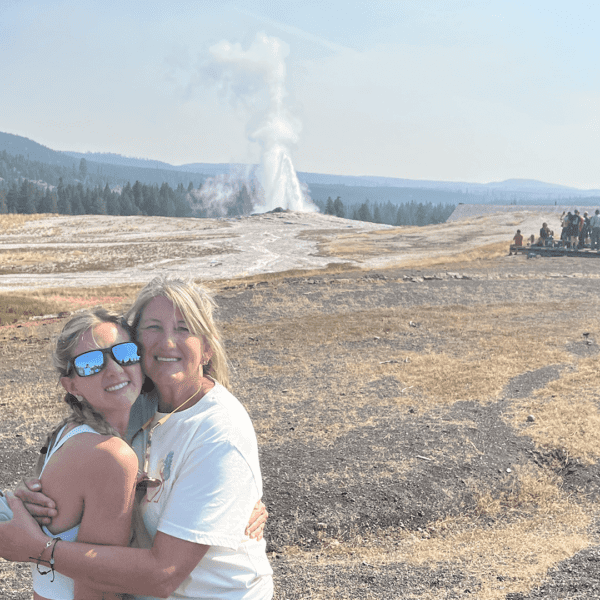 A mother and daughter pose together for a photo as Old Faithful geyser erupts in the background. Taken on a Teton Excursions tour of Yellowstone National Park.
